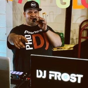 May include: A man wearing a black t-shirt with the text "Pioneer Pro DJ" in white and red, a black baseball cap with the text "RCF" in white, and a microphone in his right hand. He is standing in front of a black laptop with the text "DJ Frost" in white.