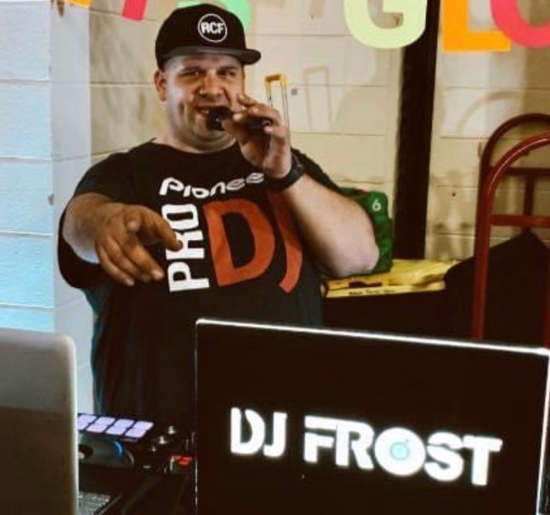 May include: A man wearing a black t-shirt with the text "Pioneer Pro DJ" in white and red, a black baseball cap with the text "RCF" in white, and a microphone in his right hand. He is standing in front of a black laptop with the text "DJ Frost" in white.