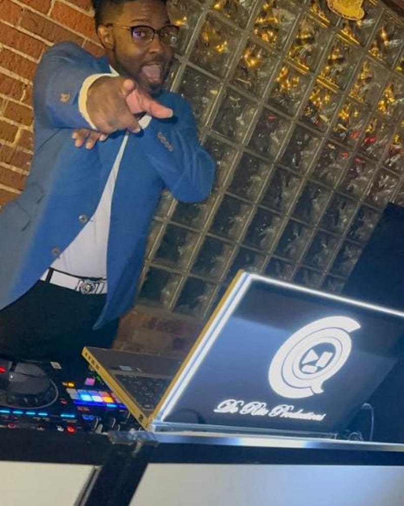 May include: A man in a blue suit is standing behind a DJ setup. He is pointing at the camera with his right hand. The DJ setup includes a laptop with a white logo that says "DJ Blue Productions" and a DJ mixer.