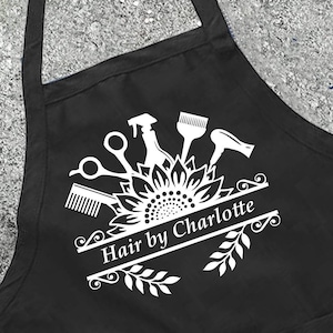 May include: Black apron with a white graphic of hairstyling tools, including scissors, a comb, and a hairdryer, above the text "Hair by Charlotte." The apron has a pocket and a neck strap.