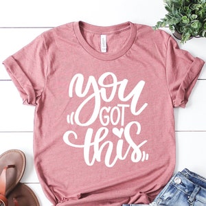 Peut inclure: Un t-shirt rose avec le texte "you got this" en lettres blanches avec un petit cœur.
