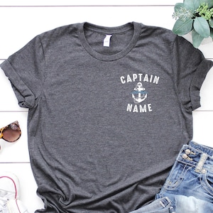 May include: A dark gray t-shirt with a white anchor and the text "CAPTAIN NAME" on the front. The shirt is laid out on a white wooden surface with a pair of sunglasses, a pair of white sneakers, and a pair of blue jeans.