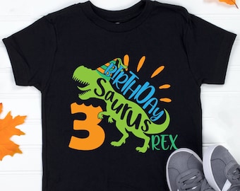 May include: Black t-shirt with a green dinosaur wearing a party hat and the text "Birthday Saurus Rex" and the number "3" in orange.
