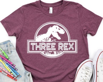 Può includere: Maglietta a maniche corte color borgogna con una grafica bianca di uno scheletro di dinosauro in un cerchio con il testo "THREE REX" in un rettangolo bianco.