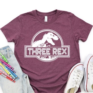 Peut inclure: T-shirt à manches courtes bordeaux avec un motif blanc d'un squelette de dinosaure dans un cercle avec le texte "THREE REX" dans un rectangle blanc.