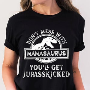 May include: Black t-shirt with white text and a dinosaur graphic. The text reads "DON'T MESS WITH MAMASAURUS YOU'LL GET JURASSKICKED" in a stylized font. The graphic features a dinosaur skeleton inside a circular emblem.