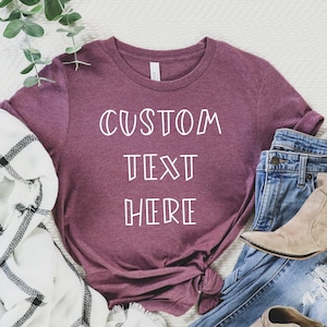 Pode incluir: Uma camiseta de cor vinho com o texto "CUSTOM TEXT HERE" em branco. A camiseta est&aacute; colocada sobre uma superf&iacute;cie branca com uma echarpe xadrez branca e preta, cal&ccedil;as de ganga azuis e botas de tornozelo castanhas.