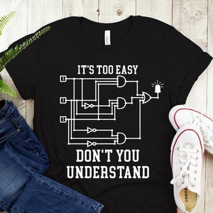 May include: Black t-shirt with white text and a circuit diagram. The text reads "IT'S TOO EASY DON'T YOU UNDERSTAND". The shirt is paired with blue jeans and white sneakers with red trim. A green plant is in the background.
