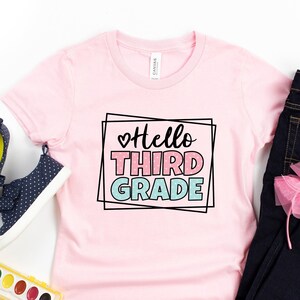 May include: Pink t-shirt with the text "Hello Third Grade" in a black frame with a pink heart. The shirt is laid out on a white surface with a pair of blue sneakers, a set of watercolors, a paintbrush, a headband with a pink bow, and a pair of blue jeans.