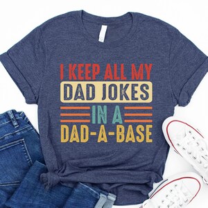 May include: A navy blue t-shirt with a vintage-style graphic that reads "I keep all my dad jokes in a dad-a-base".