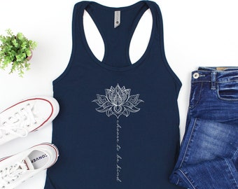May include: Navy blue racerback tank top with a white lotus flower design and the words "choose to be kind". The tank top is paired with blue jeans and white sneakers.