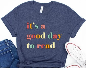 May include: A navy blue t-shirt with the text "it's a good day to read" in colourful letters. The shirt is paired with blue jeans and white trainers.