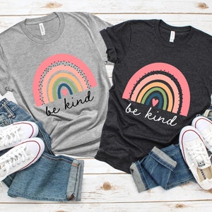 May include: Two t-shirts with a rainbow and the text "be kind" printed on them. The shirts are gray and black and are laid out on a white wooden surface with jeans and white sneakers.