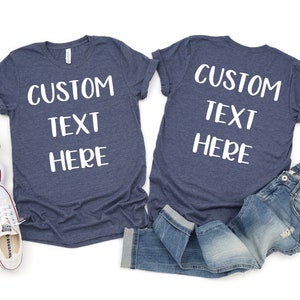 Op de afbeelding: Twee marineblauwe T-shirts met witte tekst die "CUSTOM TEXT HERE" zegt. Een shirt ligt plat en het andere is gevouwen en ligt op een blauwe spijkerbroek.