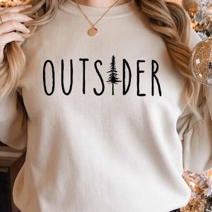 Peut inclure: Un sweat-shirt crème avec le texte "OUTSIDER" imprimé en noir. Un petit graphique d'arbre de pin est centré entre les mots.