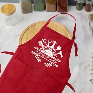 May include: A red apron with two front pockets and a neck strap. The apron features a white graphic of hairstyling tools and the text "Hair by Charlotte". The apron is designed for hairstylists.