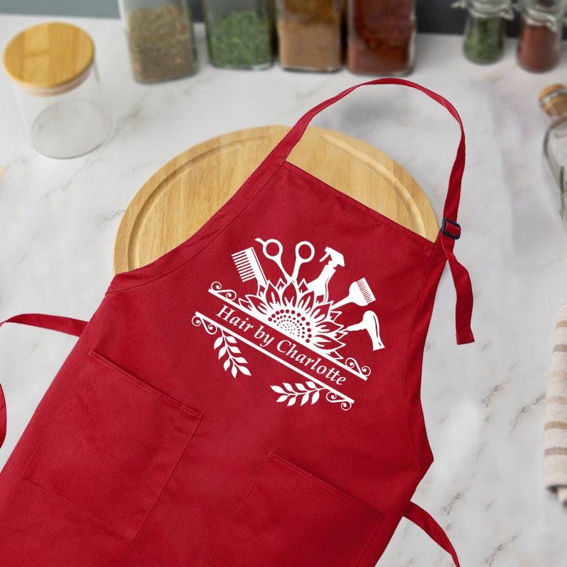 May include: A red apron with two front pockets and a neck strap. The apron features a white graphic of hairstyling tools and the text "Hair by Charlotte". The apron is designed for hairstylists.