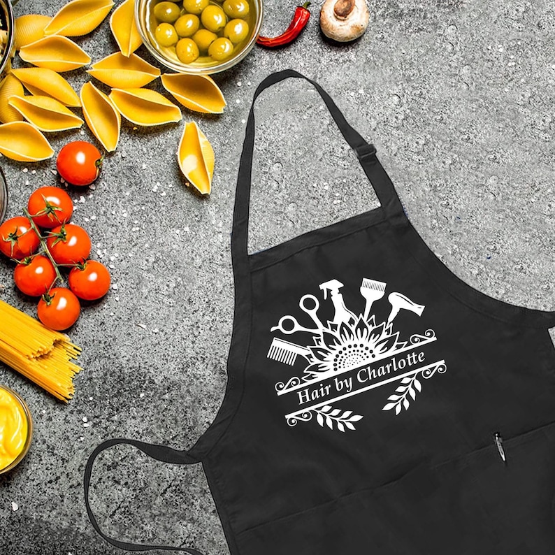 May include: Black apron with a white graphic of hairstyling tools, including scissors, a comb, and a hairdryer, above the text "Hair by Charlotte." The apron has a pocket and a neck strap.
