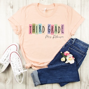Op de afbeelding: Een perzikkleurig T-shirt met de tekst "THIRD GRADE" in kleurrijke blokletters en "Mrs. Robinson" in zwarte cursieve letters eronder. Het shirt ligt op een wit houten oppervlak met een paar witte sneakers en een blauwe jeans.