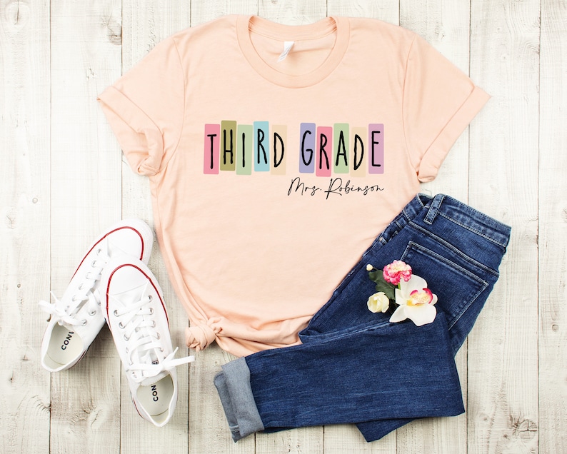 Op de afbeelding: Een perzikkleurig T-shirt met de tekst "THIRD GRADE" in kleurrijke blokletters en "Mrs. Robinson" in zwarte cursieve letters eronder. Het shirt ligt op een wit houten oppervlak met een paar witte sneakers en een blauwe jeans.