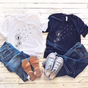 Puede incluir: Dos camisetas con un dise&ntilde;o de diente de le&oacute;n. La camiseta blanca tiene un dise&ntilde;o de diente de le&oacute;n negro y la camiseta negra tiene un dise&ntilde;o de diente de le&oacute;n blanco. Ambas camisetas est&aacute;n dispuestas sobre una superficie de madera blanca con pantalones vaqueros azules y sandalias marrones.