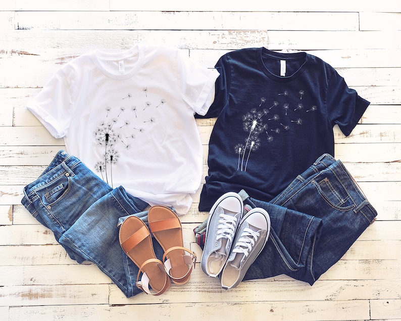 Puede incluir: Dos camisetas con un dise&ntilde;o de diente de le&oacute;n. La camiseta blanca tiene un dise&ntilde;o de diente de le&oacute;n negro y la camiseta negra tiene un dise&ntilde;o de diente de le&oacute;n blanco. Ambas camisetas est&aacute;n dispuestas sobre una superficie de madera blanca con pantalones vaqueros azules y sandalias marrones.