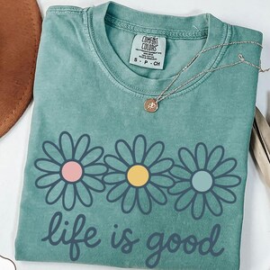 a green t-shirt with a flower design and the text "life is good" printed on it, along with a necklace and a hat.