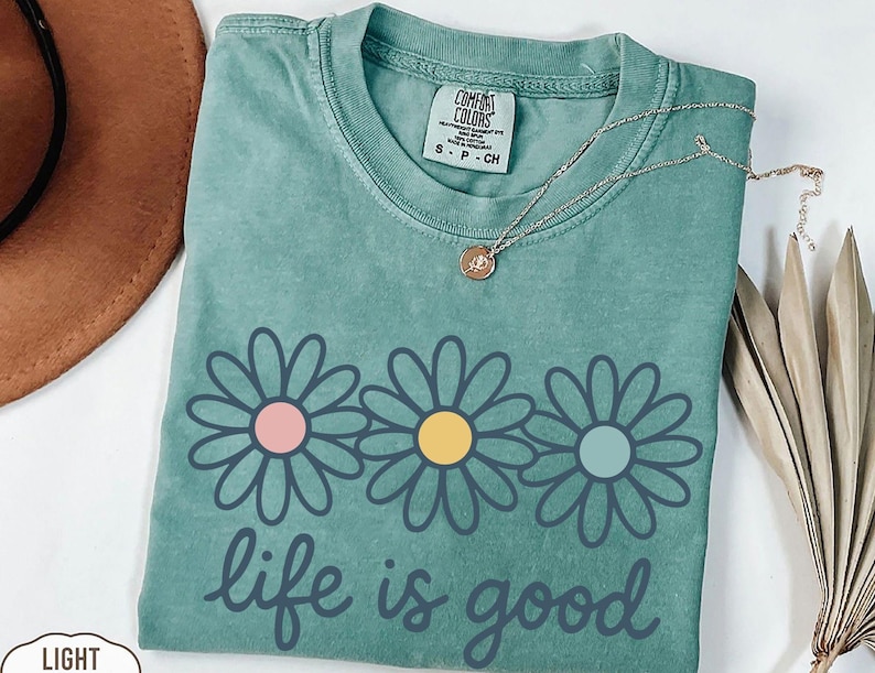 a green t-shirt with a flower design and the text "life is good" printed on it, along with a necklace and a hat.