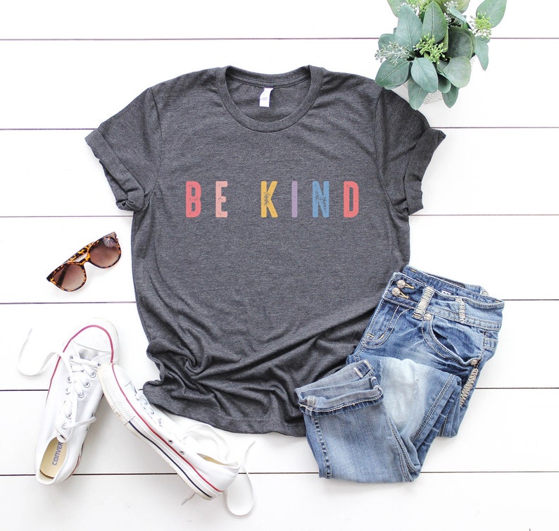 May include: A dark gray t-shirt with the words "BE KIND" printed in colorful letters. The letters are in shades of pink, orange, yellow, blue, and red. The shirt is laid flat on a white wooden surface with a pair of white sneakers, blue jeans, and sunglasses.