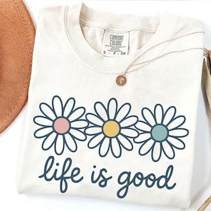 a white t-shirt with a graphic design of three colorful flowers and the text "life is good" printed on it. The t-shirt is placed on a white surface, along with a brown hat, a necklace, and some dried leaves.