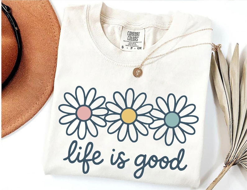 a white t-shirt with a graphic design of three colorful flowers and the text "life is good" printed on it. The t-shirt is placed on a white surface, along with a brown hat, a necklace, and some dried leaves.