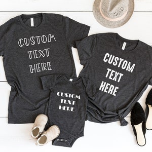 Pode incluir: Tr&ecirc;s camisetas cinzas com o texto "CUSTOM TEXT HERE" em branco. As camisetas est&atilde;o dispostas sobre uma superf&iacute;cie de madeira branca com um chap&eacute;u, &oacute;culos de sol e sapatos.