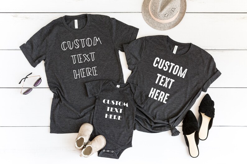 Pode incluir: Tr&ecirc;s camisetas cinzas com o texto "CUSTOM TEXT HERE" em branco. As camisetas est&atilde;o dispostas sobre uma superf&iacute;cie de madeira branca com um chap&eacute;u, &oacute;culos de sol e sapatos.