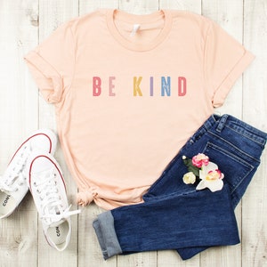 May include: A peach-colored t-shirt with the text "BE KIND" in rainbow colors. The shirt is laid out on a white wooden surface with a pair of white sneakers and blue jeans with flowers.