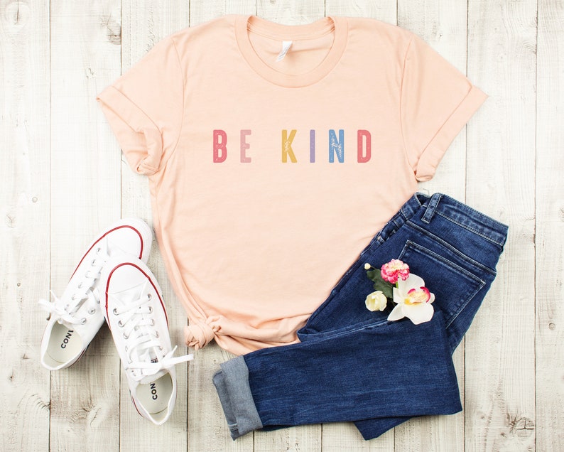 May include: A peach-colored t-shirt with the text "BE KIND" in rainbow colors. The shirt is laid out on a white wooden surface with a pair of white sneakers and blue jeans with flowers.