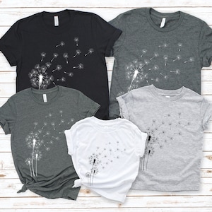 Puede incluir: Cuatro camisetas con un dise&ntilde;o de diente de le&oacute;n. Las camisetas son de color negro, gris oscuro, gris claro y blanco.