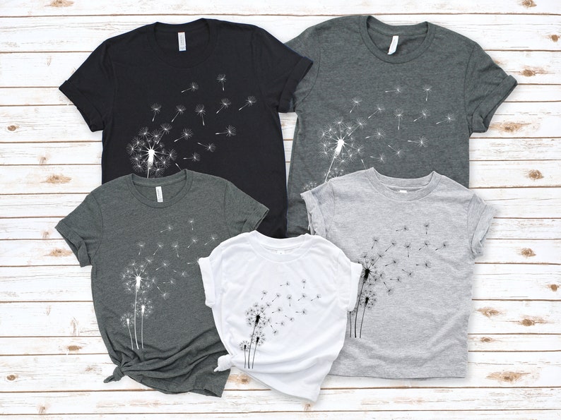 Puede incluir: Cuatro camisetas con un dise&ntilde;o de diente de le&oacute;n. Las camisetas son de color negro, gris oscuro, gris claro y blanco.