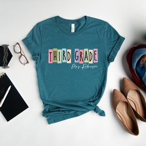 Op de afbeelding: Tealblauw T-shirt met de tekst "THIRD GRADE" in kleurrijke blokletters en "Mrs. Robinson" in wit handschrift eronder.