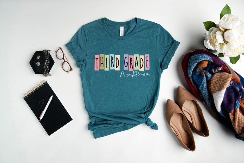 Op de afbeelding: Tealblauw T-shirt met de tekst "THIRD GRADE" in kleurrijke blokletters en "Mrs. Robinson" in wit handschrift eronder.