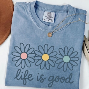 a blue t-shirt with a flower design and the text "life is good" printed on it, along with a necklace and a hat.