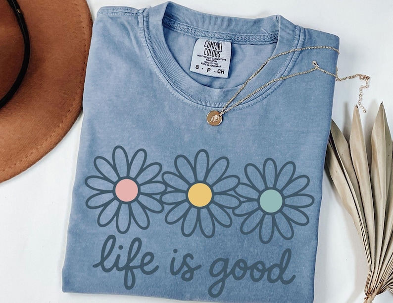 a blue t-shirt with a flower design and the text "life is good" printed on it, along with a necklace and a hat.
