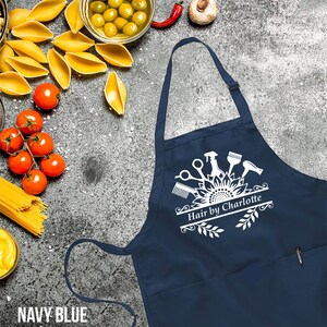 May include: Navy blue apron with a white graphic featuring scissors, a comb, a hair dryer, and the text "Hair by Charlotte." The apron has a pocket and adjustable neck strap. The words "NAVY BLUE" are at the bottom.