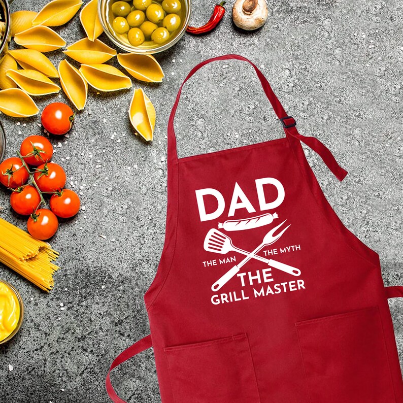 May include: Red apron with white text that reads "Dad The Man The Myth The Grill Master" with a grill spatula and fork graphic.
