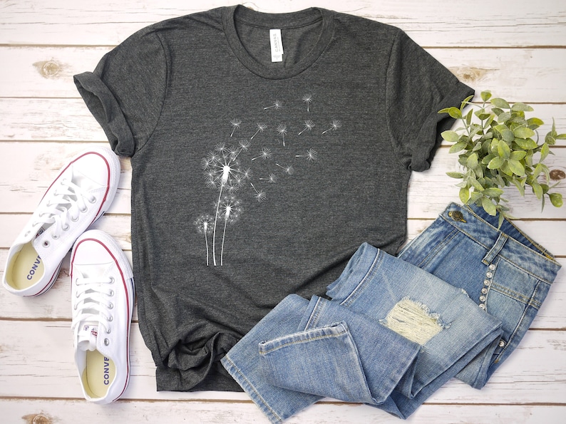 Puede incluir: Una camiseta gris oscuro con un dise&ntilde;o de diente de le&oacute;n blanco. El diente de le&oacute;n est&aacute; soplando semillas en el viento. La camiseta est&aacute; colocada sobre una superficie de madera blanca con un par de zapatillas blancas y unos vaqueros azules.