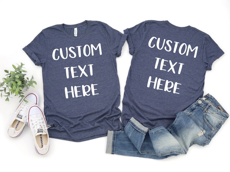 Op de afbeelding: Twee marineblauwe T-shirts met witte tekst die "CUSTOM TEXT HERE" zegt. Een shirt ligt plat en het andere is gevouwen en ligt op een blauwe spijkerbroek.