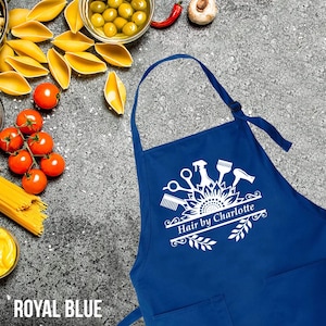 May include: Royal blue apron with a white graphic of salon tools and the text "Hair by Charlotte." The apron has a neck strap and two front pockets. The apron is displayed on a textured gray surface.