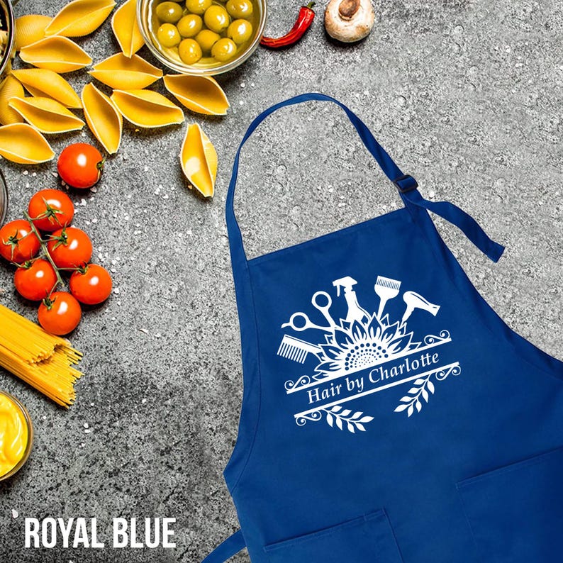 May include: Royal blue apron with a white graphic of salon tools and the text "Hair by Charlotte." The apron has a neck strap and two front pockets. The apron is displayed on a textured gray surface.