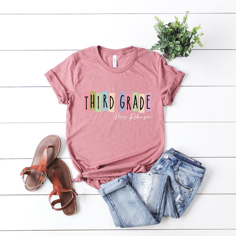Op de afbeelding: Een roze T-shirt met de tekst "THIRD GRADE" in kleurrijke blokken en "Mrs. Robinson" in zwarte tekst eronder.