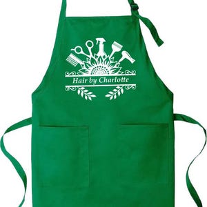 May include: Green apron with two front pockets and a neck strap. The apron features a white graphic of salon tools and the text "Hair by Charlotte". The design includes scissors, a comb, a spray bottle, and a hairdryer.
