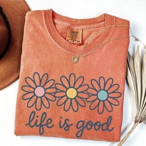 a red t-shirt with a flower design and the text "life is good" printed on it, along with a brown hat and a fan.